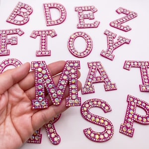 May include: Pink felt letters covered in clear rhinestones. The letters are in uppercase and include the letters B, D, E, F, O, T, Y, Z, M, A, S, P, R.