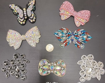 Puede incluir: Surtido de adornos de pedrería, que incluyen una mariposa, lazos y racimos de pedrería. Los adornos son de varios colores, entre ellos rosa, blanco, azul, rojo y plateado.