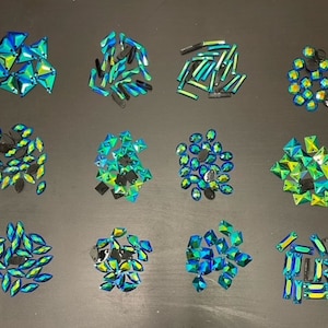 以下が含まれることがあります： 緑と黒の虹色のラインストーンが、様々な形とサイズで詰め合わせられています。ラインストーンは、黒い表面に10つのグループに分けて配置されています。