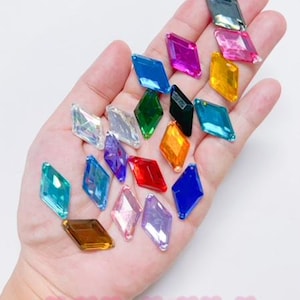 May include: Assortment of colourful diamond-shaped rhinestones in various colours, including blue, green, red, pink, orange, and clear. The rhinestones are arranged on a hand.