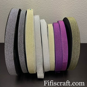 May include: Assortment of elastic trim with glitter in various colors, including black, silver, gold, and purple. The trim is rolled up and stacked on a table.