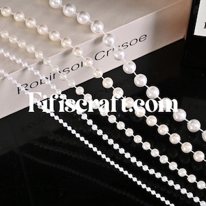 May include: Five strands of white pearl bead garland, each with different sized beads, arranged in a fan shape on a white surface. The text "Robinson Crusoe" is visible on the surface.