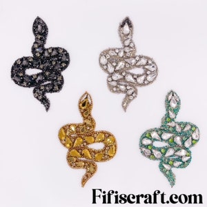 May include: Four bejeweled snake appliques in black, silver, gold, and green. Each snake is intricately detailed with various sizes of sparkling crystals and beads. The appliques are arranged on a white background, with the website address "Fifiscraft.com" at the bottom.