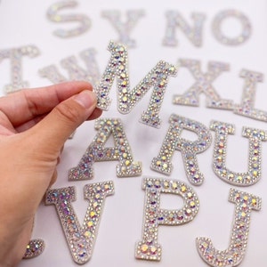 May include: A set of iron-on rhinestone letters in various sizes. The letters are made of white fabric with clear iridescent rhinestones. The letters are arranged in a random order on a white surface.