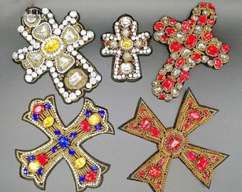 May include: Five different cross-shaped embellishments with gold, red, blue, and clear rhinestones. The crosses are made of fabric and are decorated with beads and sequins.
