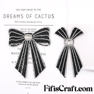 May include: Two black and white bow-shaped fabric patches with rhinestones and a clear crystal in the center. The text "THE EASY GUIDE TO THE DREAMS OF CACTUS" is at the top of the image. The text "With succulent specialist Jon Green" is below the first line of text. The text "TEXT BY JON GREEN" is above the bows. The text "RAPHY BY FORTY" is below the first line of text. The text "To live simply is my only wish, nature as my best friend" is below the bows. The text "FifisCraft.com" is at the bottom of the image.