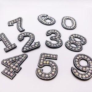 May include: Ten black iron-on patches with numbers 0-9, each number is covered in iridescent rhinestones.