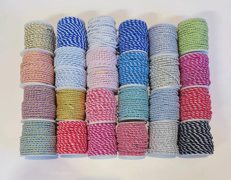 May include: A collection of twenty-four spools of twisted cord arranged in a grid. The cords feature various colour combinations including solid metallic gold, silver, neon green, and multi-coloured striped patterns in blue, pink, red, and black.