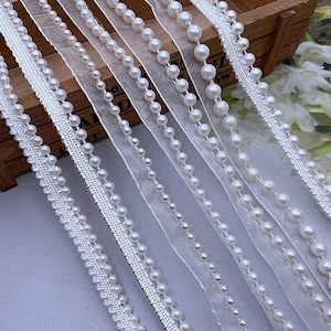 May include: A collection of white pearl trim with varying pearl sizes and styles. The trim is on a white mesh or sheer fabric background.