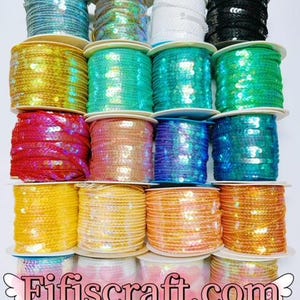 Può includere: Una collezione di rocchetti di passamaneria di paillettes in vari colori, tra cui blu, verde, oro, rosso e arancione. I rocchetti sono impilati. Il testo "Fifiscraft.com" è visibile in basso.