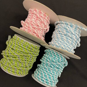 May include: Four spools of twisted cord in distinct colour combinations. One spool features pink and white, another light blue and white, a third lime green and silver, and the fourth turquoise and white. Each cord is wound around a circular spool.