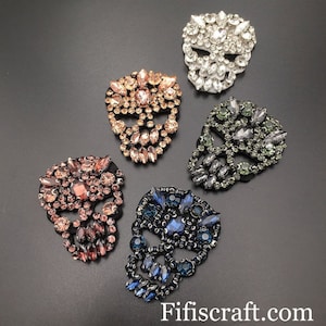 Può includere: Cinque decorazioni a forma di teschio con strass di diversi colori. Gli strass sono trasparenti, rosa, blu, verdi e neri.