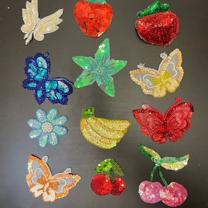 Puede incluir: Surtido de 12 parches termoadhesivos coloridos con varios diseños, incluyendo mariposas, flores, frutas y una estrella de mar. Los parches están hechos de lentejuelas y cuentas.