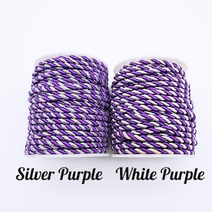 May include: Two spools of twisted cord. The left spool features a silver and purple twisted cord labelled Silver Purple. The right spool features a white and purple twisted cord labelled White Purple. Both cords are wound tightly around white spools.