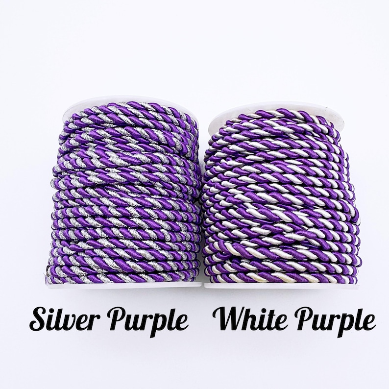 May include: Two spools of twisted cord. The left spool features a silver and purple twisted cord labelled Silver Purple. The right spool features a white and purple twisted cord labelled White Purple. Both cords are wound tightly around white spools.