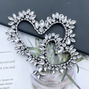 Puede incluir: Un broche plateado en forma de corazón adornado con numerosos cristales transparentes y brillantes. El broche presenta dos contornos de corazón entrelazados, cada uno adornado con cristales de varios tamaños y formas, creando un efecto deslumbrante. El diseño está sobre un fondo oscuro.