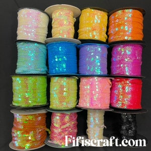 Puede incluir: Una colección de carretes de cinta de lentejuelas en varios colores, incluyendo blanco, amarillo, naranja, azul, rosa, verde y negro. La cinta está enrollada en carretes blancos y negros. El sitio web "Fifiscraft.com" es visible en la parte inferior.