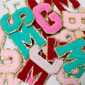 May include: A collection of colorful felt letters with gold glitter trim. The letters are in various shades of pink, red, turquoise, and white.
