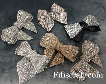 Puede incluir: Seis lazos adornados con pedrería en varios colores, incluyendo plata, oro e iridiscente. Los lazos están dispuestos sobre una superficie gris con el texto "Fifiscraft.com" visible en la esquina inferior derecha.