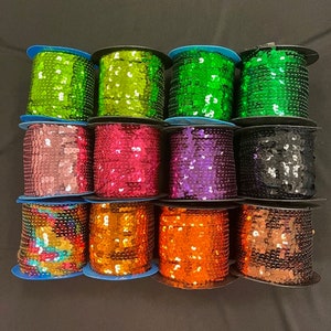 May include: Twelve spools of sequined trim in various colors, including green, pink, purple, black, orange, and iridescent. The spools are arranged in three rows of four.