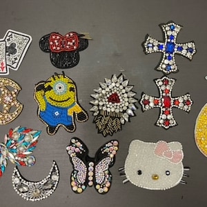 May include: An assortment of iron-on patches featuring various designs, including a pineapple, a cross, a butterfly, a crescent moon, a minion, a playing card, a Mickey Mouse head, and a Hello Kitty head. The patches are decorated with colourful rhinestones and beads.