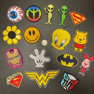 Può includere: Una collezione di toppe da stirare con vari disegni, tra cui un segno di pace, un viso sorridente, un uccello dei cartoni animati, un logo di supereroe e un alieno dei cartoni animati. Le toppe sono colorate e hanno una varietà di forme e dimensioni.