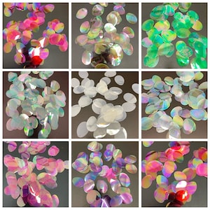 May include: Assortment of iridescent oval sequins in various colors, including green, white, pink, and purple. The sequins are scattered on a black surface.
