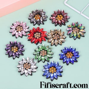 May include: Assortment of ten colorful flower-shaped embellishments with rhinestones. Each flower has a different color and is decorated with clear and gold rhinestones.