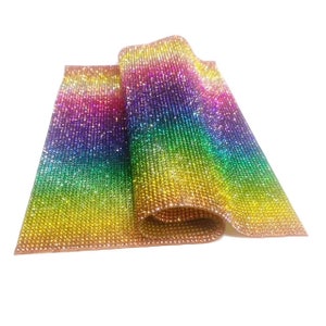 Pode incluir: Uma folha de tecido de strass arco-íris. O tecido é feito de pequenos strass iridescentes que são dispostos em um padrão de degradê. O tecido está dobrado ao meio e os strass brilham na luz.
