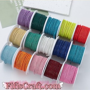 Könnte beinhalten: Sortiment an bunten Strass-Zierbändern auf Spulen. Das Band besteht aus kleinen quadratischen Strasssteinen in verschiedenen Farben, darunter Lila, Grün, Blau, Gelb, Rot, Weiß, Orange, Rosa, Burgunderrot und Hellrosa. Die Spulen sind in einem Halbkreis auf einer weißen Oberfläche angeordnet.