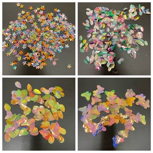 May include: Assortment of iridescent sequins in various shapes, including flowers, butterflies, and teardrop shapes. The sequins are scattered on a black background.