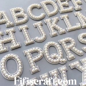 Könnte beinhalten: Ein Satz weißer Perlen- und Strass-Buchstaben. Die Buchstaben sind in Großbuchstaben und alphabetisch geordnet. Die Buchstaben befinden sich auf einem weißen Hintergrund.