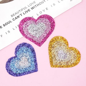 以下が含まれることがあります： キラキラと輝くラインストーンのデザインが施された、ハート型のアイロン接着パッチが3つ。ハートはピンク、ブルー、ゴールドです。