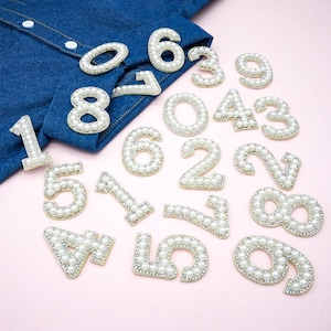May include: A set of pearl and rhinestone number appliques in white. The numbers are arranged on a pink surface.