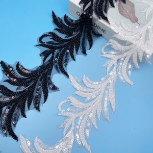 Könnte beinhalten: Zwei Borten aus paillettenbesetztem Stoff, eine schwarze und eine weiße, mit einem federartigen Design. Der Stoff liegt auf einem blauen Hintergrund.