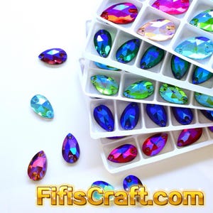 May include: A collection of teardrop-shaped faceted crystals in various iridescent colors, including blue, green, pink, and purple, arranged in a white plastic sorting tray with several loose crystals scattered nearby. The text FifisCraft.com is visible.