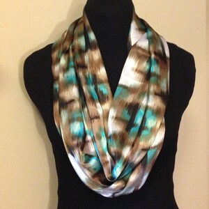 May include: An infinity scarf with a colorful abstract pattern. The scarf features a mix of teal, brown, and white hues. The fabric appears to be silky and drapes in soft folds. The scarf is displayed on a black mannequin.