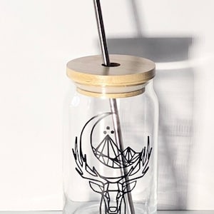 Puede incluir: Un tarro de vidrio transparente con tapa de madera y una pajita de acero inoxidable. El tarro tiene un diseño geométrico en blanco y negro de un ciervo con una luna creciente y montañas.