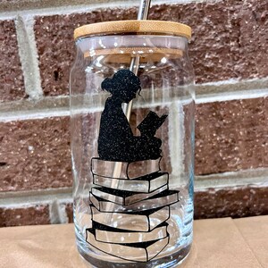 May include: A clear glass drinking cup with a bamboo lid and a silver straw. The cup features a black silhouette of a person reading on top of a stack of books. The design is made with glitter.