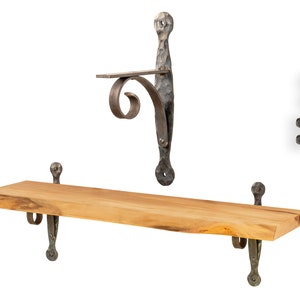 Może przedstawiać: Drewniana półka z dwoma metalowymi wspornikami. Półka wykonana jest z naturalnego drewna z żywym krawędzią. Wsporniki są ciemnobrązowe i mają ozdobny wzór w postaci zwijanego ornamentu. Półka jest zamontowana na białej ścianie.