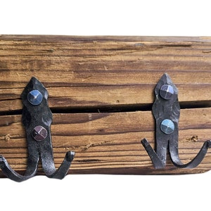 Pode incluir: Um gancho de parede de madeira rústica com dois ganchos de metal preto. Os ganchos têm detalhes decorativos e são fixados à madeira com parafusos.