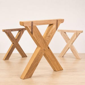 Könnte beinhalten: Drei Tischbeine aus Holz mit X-förmigem Design. Die Beine sind aus Naturholz in drei verschiedenen Farbtönen gefertigt: ein dunkles Braun, ein mittleres Eichengold und ein helles, blondes Holz. Jedes Bein hat eine horizontale obere Leiste.