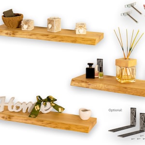 Op de afbeelding: Drie houten planken met natuurlijke randen, elk met verschillende decoratieve items. De bovenste plank heeft een rieten mand, drie kleine witte doosjes met harten en een klein wit doosje met een hart. De middelste plank heeft een glazen fles met een houten deksel, een doorzichtige glazen fles met een houten deksel en een klein flesje parfum. De onderste plank heeft een wit houten bord met de tekst "Home" met een groen lint en een kleine witte keramische pot. De planken zijn met metalen beugels aan de muur bevestigd. Optionele beugels worden ook op de afbeelding getoond.