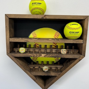 Puede incluir: Una vitrina de madera con forma de diamante de béisbol con dos pelotas de softball amarillas y dos anillos de campeonato. La vitrina tiene un fondo negro con un gráfico de pelota de softball amarilla.