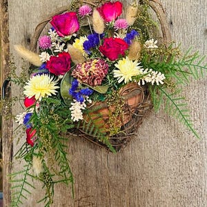 May include: A heart-shaped woven basket filled with a variety of dried flowers. The arrangement includes vibrant red roses, yellow daisies, and blue statice, accented with green foliage and tan grasses. The basket is suspended against a weathered wooden background.