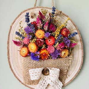 May include: A circular wood slice wall hanging featuring a burlap pocket filled with dried flowers in shades of orange, pink, purple, and yellow. The pocket is decorated with a white lace ribbon and a brown button. A jute rope is attached for hanging.