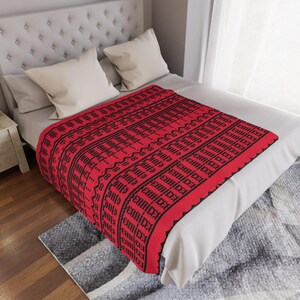 May include: A red blanket with a black geometric pattern and text. The blanket is folded over a bed with white pillows and a headboard. The blanket's design features repeating patterns and text.
