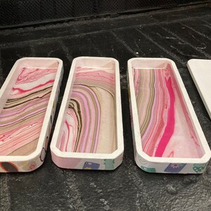May include: Three rectangular wooden trays with white rims and colorful marbled interiors. The trays have a decorative patterned exterior. A white lid with a small knob is next to the trays. The trays are ideal for organizing small items.