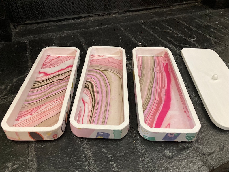 May include: Three rectangular wooden trays with white rims and colorful marbled interiors. The trays have a decorative patterned exterior. A white lid with a small knob is next to the trays. The trays are ideal for organizing small items.