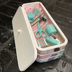 May include: A white, rectangular box with a hinged lid, containing various teal-colored office supplies. The box is decorated with a patterned design and includes scissors, tape dispenser, stapler, and paper clips.
